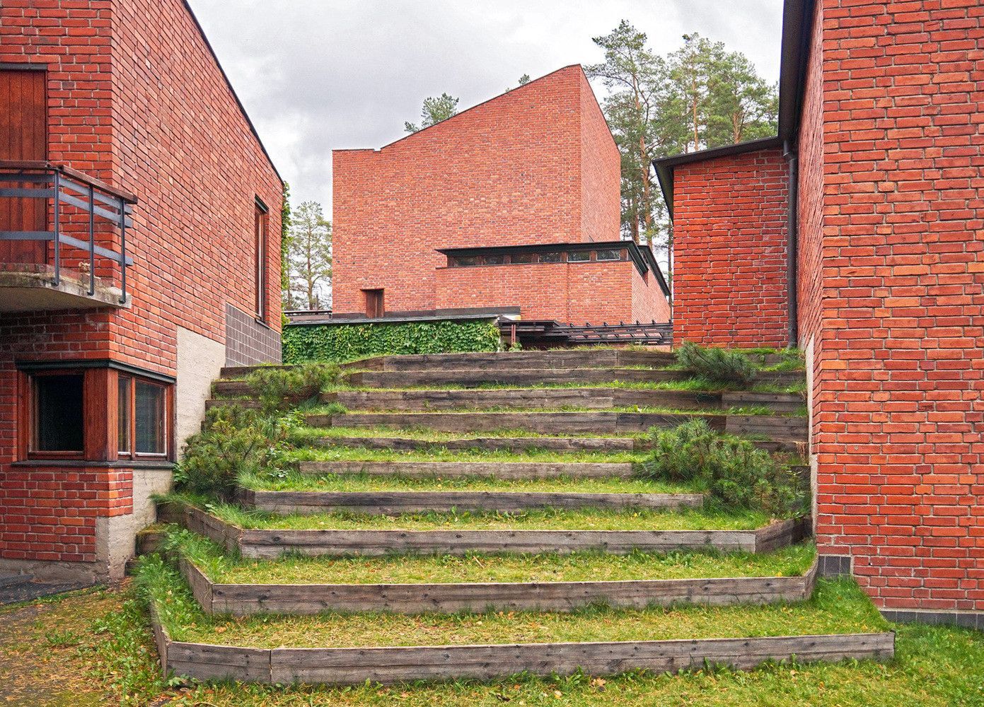 Detalle del ayuntamiento de Säynätsalo, Finlandia. Obra de Alvar Aalto. Se observa una escalera exterior realizada únicamente con listones de madera para escalonar el terreno, en el que crece la vegetación espontánea. A los lados,  y de fondo, las paredes del edificio de formas simples y geométricas, en ladrillo visto y con pequeñas ventanas y puertas de madera. Una celosía en el módulo del fondo permite la entrada de luz natural tan característica de las obras de Aalto. © Wikipedia: Kulmalukko CC BY-SA 3.0