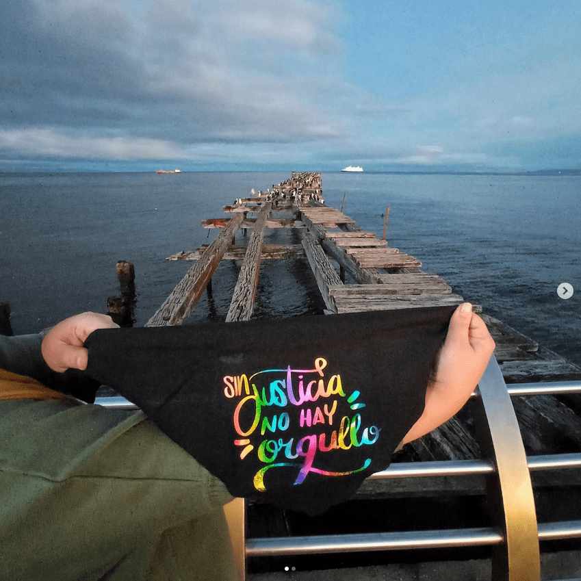 Fotografia de una pañoleta estampada en vinilo textil con un leterring que dice ¨Sin Justicia No Hay Orgullo" la pañoleta es sostenida por dos manos y de fondo se ve un muelle en el estrecho de Magallanes.