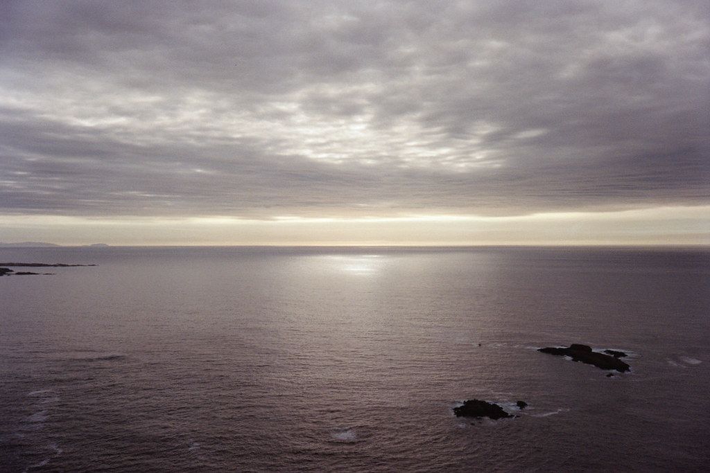 A landscape depicting a sunset over the Atlantic, featuring clouds and the sea, some rocks and the sun hidden behind the clouds.